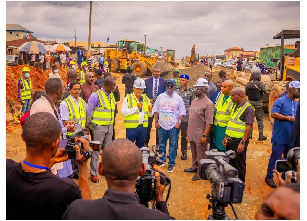 A large group of officials, engineers and press gathered on an active construction front, with earthmoving plant working behind them.